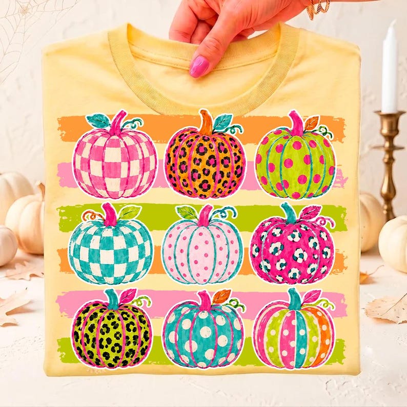 May include: A pale yellow t-shirt featuring a graphic print of nine pumpkins arranged in three rows. Each pumpkin displays a unique pattern, including checkered, leopard print, and polka dot designs in vibrant shades of pink, green, and orange.