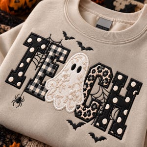 May include: A beige crewneck sweatshirt featuring the embroidered word "GHOST" with a white ghost replacing the letter "O". The letters are decorated with black and white polka dots, plaid, and floral patterns, accented by spiderwebs, bats, and a spider.