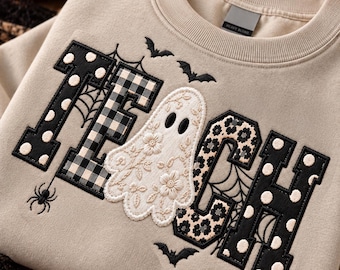 May include: A beige crewneck sweatshirt featuring the embroidered word "GHOST" with a white ghost replacing the letter "O". The letters are decorated with black and white polka dots, plaid, and floral patterns, accented by spiderwebs, bats, and a spider.