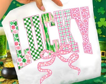 Könnte beinhalten: Weißes T-Shirt mit dem Wort "LUCKY" in einer verspielten Schriftart. Die Buchstaben weisen verschiedene Muster auf, darunter grün-weiß kariert, rosa-grün kariert und rosa Blumenmuster. Am unteren Rand befindet sich eine rosa Schleife mit Punkten.