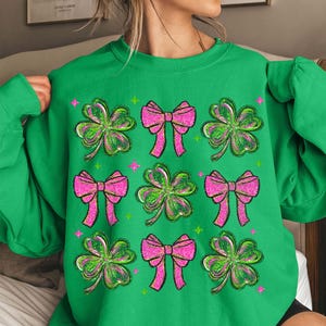 May include: Green sweatshirt featuring a pattern of pink bows and green shamrocks. The shamrocks have green and pink glitter accents. The design includes small pink and green starbursts. A festive St. Patrick's Day themed top.