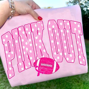 May include: A light pink sweatshirt featuring the text PINK OUT in large, bold, white-polka-dotted pink letters. Below the text is a graphic of a pink football with a white stripe and a pink awareness ribbon integrated into the design.