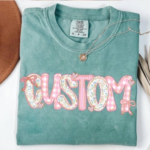 May include: A teal green t-shirt with the word "CUSTOM" in pink and white letters with a floral and gingham pattern. The letters are outlined in gold. The shirt has a tag that says "Comfort Colors" and "S - P - CH".