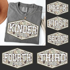 May include: A gray t-shirt with a large, distressed-style graphic that reads "KINDER GARTEN" in white letters over a camouflage pattern. Additional grade level graphics are also displayed, including "PRE-K", "FIRST GRADE", "SECOND GRADE", "FOURTH GRADE", and "THIRD GRADE".
