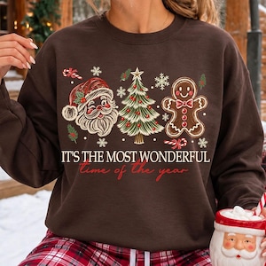 K&ouml;nnte beinhalten: Ein dunkelbraunes Sweatshirt mit Rundhalsausschnitt und einem Grafikdruck von Weihnachtsmann, geschm&uuml;cktem Weihnachtsbaum und Lebkuchenmann. Der Text lautet "IT'S THE MOST WONDERFUL time of the year", umgeben von kleinen Schneeflocken und Zuckerstangen.