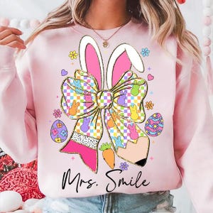 May include: Pink sweatshirt with a graphic of a bow with bunny ears, pastel colored eggs, and the text "Mrs. Smile".