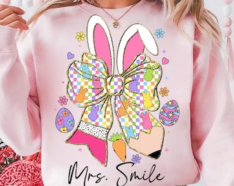 Può includere: Felpa rosa con una grafica di un fiocco con orecchie di coniglio, uova di Pasqua color pastello e il testo "Mrs. Smile".