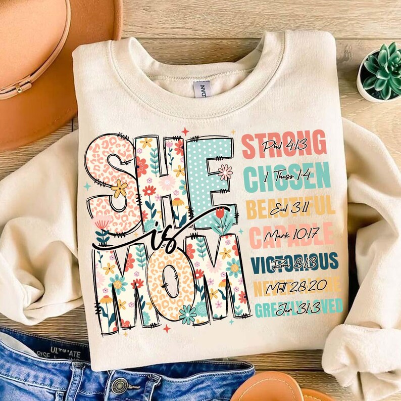 K&ouml;nnte beinhalten: Ein cremefarbener Pullover mit dem Text "She is Mom" in bunten Buchstaben mit Blumen- und Leopardenmustern. Der Text ist umgeben von einer Spalte mit W&ouml;rtern und Bibelversen, darunter "Strong", "Chosen", "Beautiful", "Capable", "Victorious", "Loved" und "Greatly Loved" mit entsprechenden Bibelzitaten.