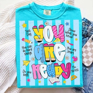 May include: A turquoise t-shirt with a colorful graphic that says "You Are Ready." The design includes school-themed elements like pencils, rulers, and stars. Additional text includes "Test Day," "You can do hard things," and "Show What you know."