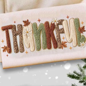 May include: A cream-coloured decorative towel embroidered with the word "THANKFUL". The letters are in various textures and colours, including leopard print, knitted patterns, and floral accents, with gold trim and autumn leaf details.
