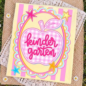 May include: A yellow canvas with pink vertical stripes and a large apple design. The apple is pink and white checkered with the words "kindergarten" in pink script. Colorful star accents complete the design.