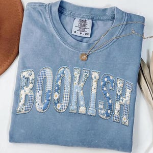 May include: A light blue Comfort Colors t-shirt with the word "BOOKISH" appliqued across the front in a patchwork design of floral and gingham patterns. A gold necklace with a pendant rests on the shirt.