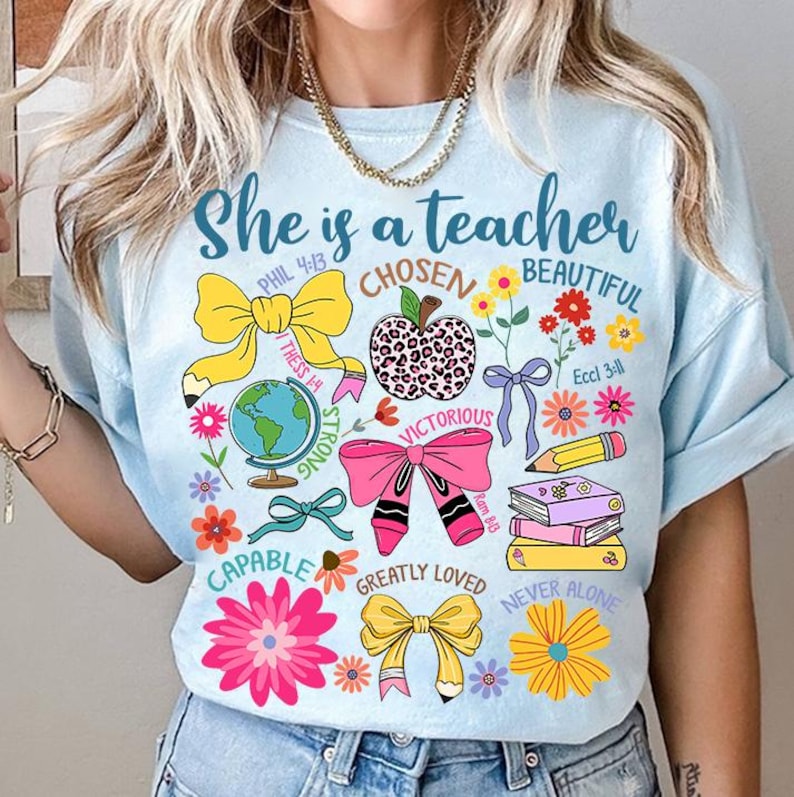 以下が含まれることがあります： 薄い青色のTシャツに「She is a teacher」という文字と、リボン、花、地球儀、本の積み重ね、リンゴなど、さまざまなカラフルなイラストが描かれています。「CHOSEN」、「BEAUTIFUL」、「STRONG」、「VICTORIOUS」、「CAPABLE」、「GREATLY LOVED」、「NEVER ALONE」という文字もTシャツに含まれています。
