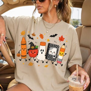 May include: A beige t-shirt featuring a graphic print of five anthropomorphic school supplies dressed for Halloween: an orange crayon, a red apple with a witch hat, a white ghost, a black notebook with a spider web, and a yellow pencil.