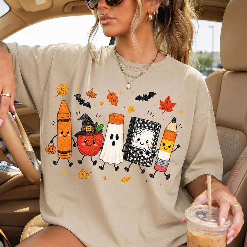 May include: A beige t-shirt featuring a graphic print of five anthropomorphic school supplies dressed for Halloween: an orange crayon, a red apple with a witch hat, a white ghost, a black notebook with a spider web, and a yellow pencil.