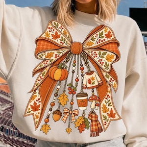 May include: A cream-colored sweatshirt featuring a large graphic of a plaid orange and brown bow. Dangling from the bow are various autumn-themed charms including a pumpkin, a sweater, a coffee cup, a mushroom, an acorn, and a scarf.