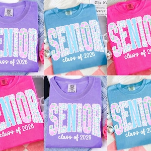 May include: A collection of six t-shirts in various colors: purple, blue, and pink. Each shirt features the word "SENIOR" in a decorative font, with "class of 2026" below. The shirts are laid out on a patterned surface.