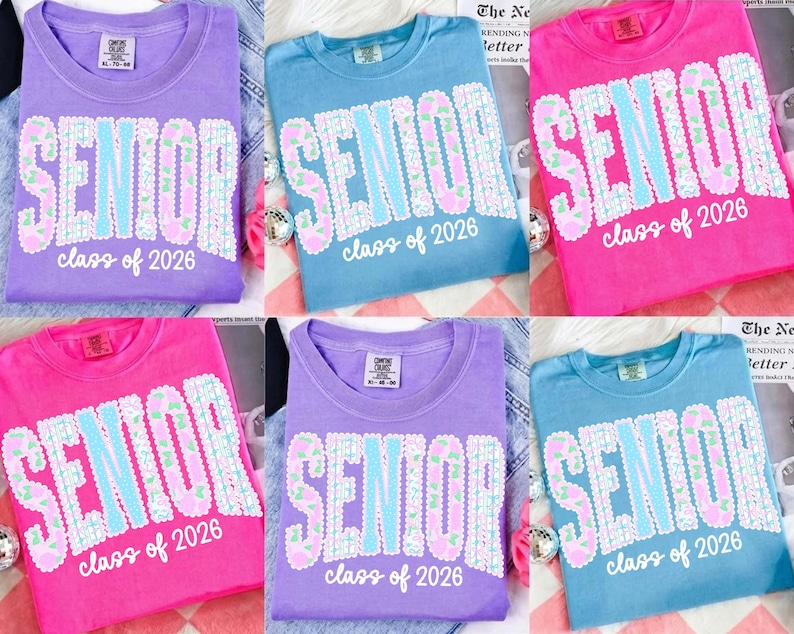 May include: A collection of six t-shirts in various colors: purple, blue, and pink. Each shirt features the word "SENIOR" in a decorative font, with "class of 2026" below. The shirts are laid out on a patterned surface.