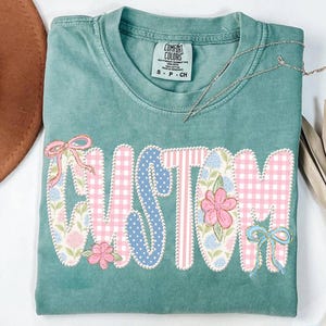 May include: A teal Comfort Colors t-shirt with the word "CUSTOM" appliqued in pink gingham, blue polka dots, and floral patterns. The design includes pink and blue bows and flowers. A silver necklace rests on the shirt.