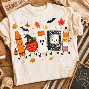 May include: A cream-colored t-shirt featuring a colorful graphic of anthropomorphic school supplies and autumn elements. The design includes a crayon, an apple wearing a witch hat, a ghost, a notebook with a spider, and a pencil, surrounded by bats and leaves.