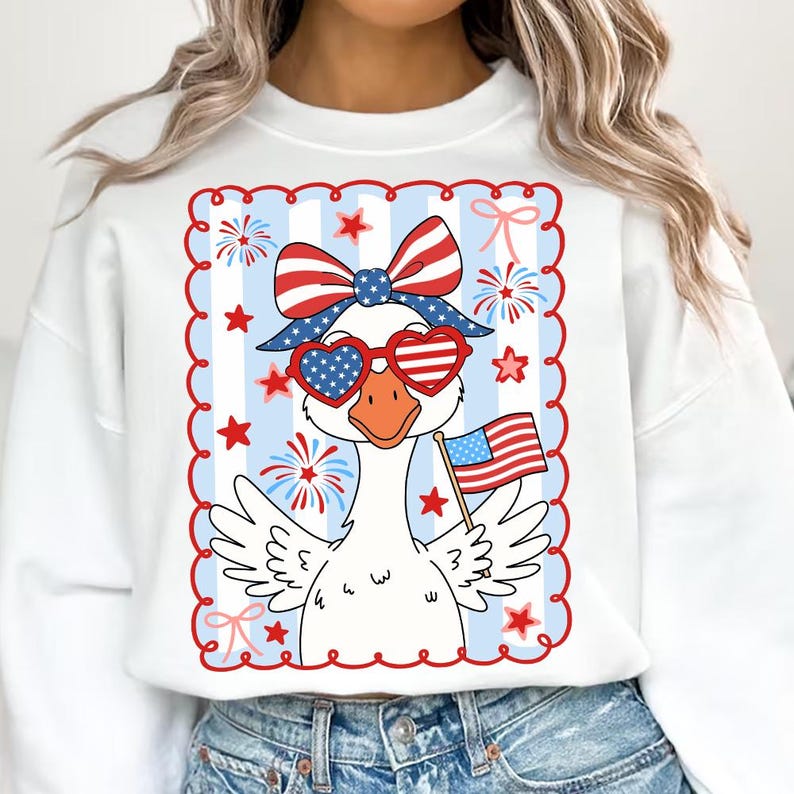 Puede incluir: Sudadera blanca con un dise&ntilde;o de ganso patri&oacute;tico con gafas de sol en forma de coraz&oacute;n y un lazo, sosteniendo una bandera estadounidense. El dise&ntilde;o incluye estrellas, fuegos artificiales y lazos rojos, blancos y azules.