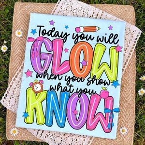 以下が含まれることがあります： 「Today you will GLOW when you show what you KNOW」というカラフルな文字がプリントされた水色のTシャツ。星、鉛筆、リンゴ、時計のイラストも含まれています。