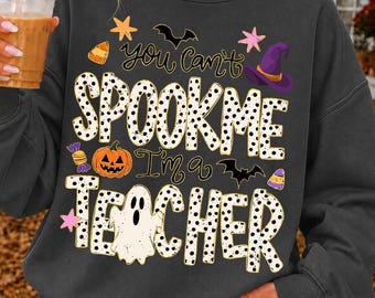 May include: A dark gray crewneck sweatshirt featuring a graphic print with the text "you can't SPOOK ME I'm a TEACHER". The design includes a white ghost, an orange pumpkin, a purple witch hat, bats, stars, and candy corn in various colors.