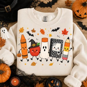 May include: A cream-colored crewneck sweatshirt featuring a graphic print of five anthropomorphic school supplies: an orange crayon, a red apple wearing a witch hat, a white ghost, a black composition notebook, and a yellow pencil, surrounded by autumn leaves and bats.