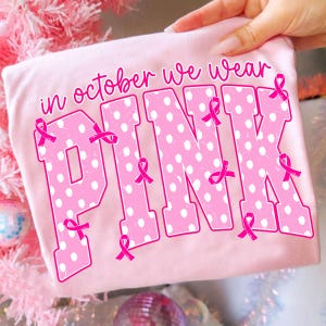 Puede incluir: Una camiseta rosa claro con el texto "in october we wear PINK" en fuente rosa oscuro. La palabra "PINK" est&aacute; escrita en letras grandes de estilo bloque con un patr&oacute;n de lunares blancos y varios lazos rosas peque&ntilde;os de concienciaci&oacute;n.