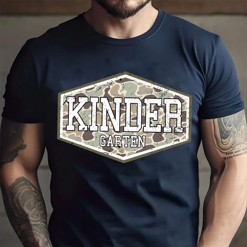 May include: Navy blue t-shirt with a distressed, camouflage-patterned hexagon graphic. The graphic features the words "KINDER" above "GARTEN" in a white, bold font, suitable for a school theme.