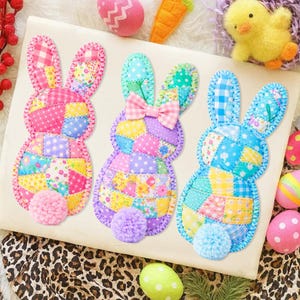 May include: Three colorful patchwork bunny decorations with pom-pom tails. Each bunny is a different color, with pink, purple, and blue accents. The bunnies are surrounded by Easter-themed decorations, including eggs and a toy chick.