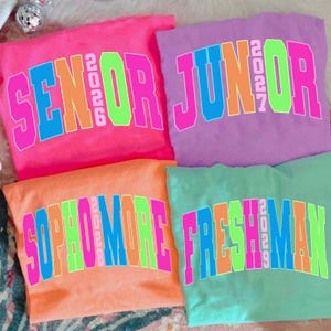 May include: Four colorful sweatshirts, each with a different class year and text. The shirts are pink, purple, orange, and green. The text on the shirts reads "SENIOR 2026", "JUNIOR 2027", "SOPHOMORE 2028", and "FRESHMAN 2029".