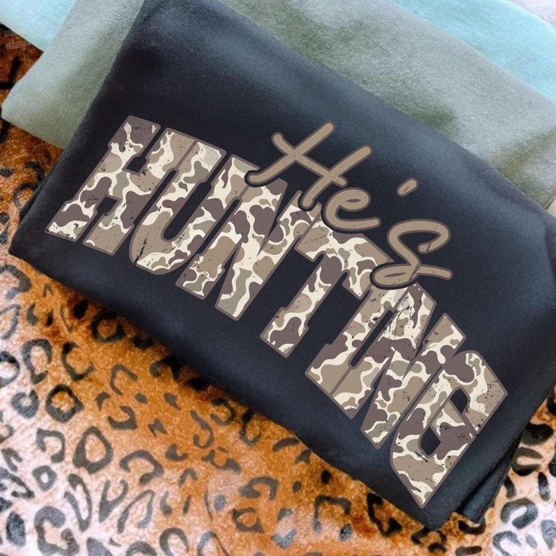 May include: Black sweatshirt with the text "He's HUNTING" in a camouflage pattern. The sweatshirt is folded, with a leopard print fabric and two solid-colored fabrics in the background.