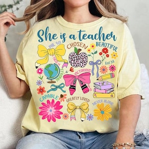 以下が含まれることがあります： 「She is a teacher」という文字と、花、リボン、地球儀、リンゴ、本、鉛筆のカラフルなデザインが施された黄色のTシャツ。デザインには聖書の言葉も含まれています。