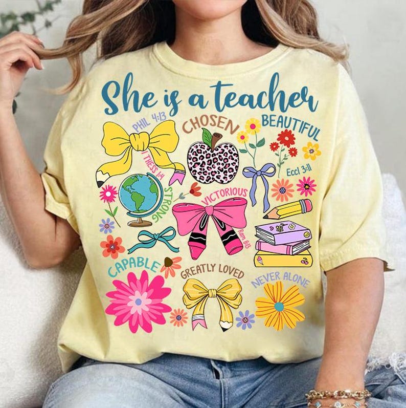 以下が含まれることがあります： 「She is a teacher」という文字と、花、リボン、地球儀、リンゴ、本、鉛筆のカラフルなデザインが施された黄色のTシャツ。デザインには聖書の言葉も含まれています。