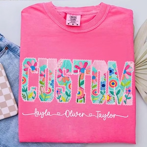 Op de afbeelding: Een felroze Comfort Colors t-shirt met het woord "CUSTOM" in een bloemen patchwork ontwerp. Onder het woord staan de namen "Layla", "Oliver" en "Taylor". Het shirt ligt op een denim oppervlak.