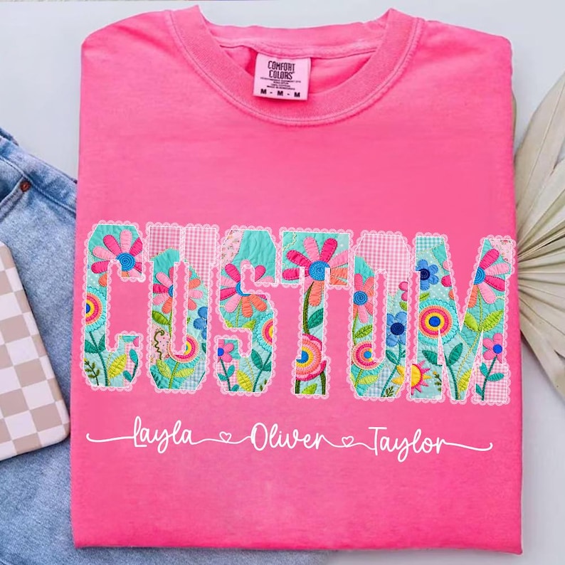 Op de afbeelding: Een felroze Comfort Colors t-shirt met het woord "CUSTOM" in een bloemen patchwork ontwerp. Onder het woord staan de namen "Layla", "Oliver" en "Taylor". Het shirt ligt op een denim oppervlak.