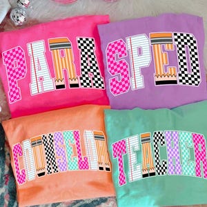 Puede incluir: Cuatro camisetas coloridas con diseños de texto. Las camisetas son rosa, morado, naranja y verde azulado. Cada camiseta tiene una palabra diferente: "PARA", "SPEED", "COUNSELOR" y "TEACHER". Las letras están diseñadas con patrones de lápiz y tablero de ajedrez.