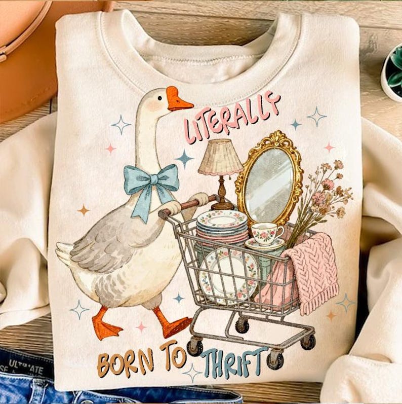 May include: Cream-colored sweatshirt with a whimsical graphic of a goose wearing a blue bow tie, pushing a shopping cart filled with vintage items. Text reads "Literally Born to Thrift."