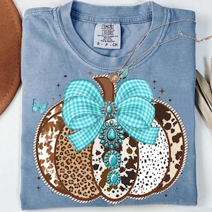 May include: A light blue Comfort Colors t-shirt featuring a pumpkin design. The pumpkin has a brown stem, a turquoise gingham bow, and a turquoise jewel design. The pumpkin is decorated with a cow print and a butterfly.