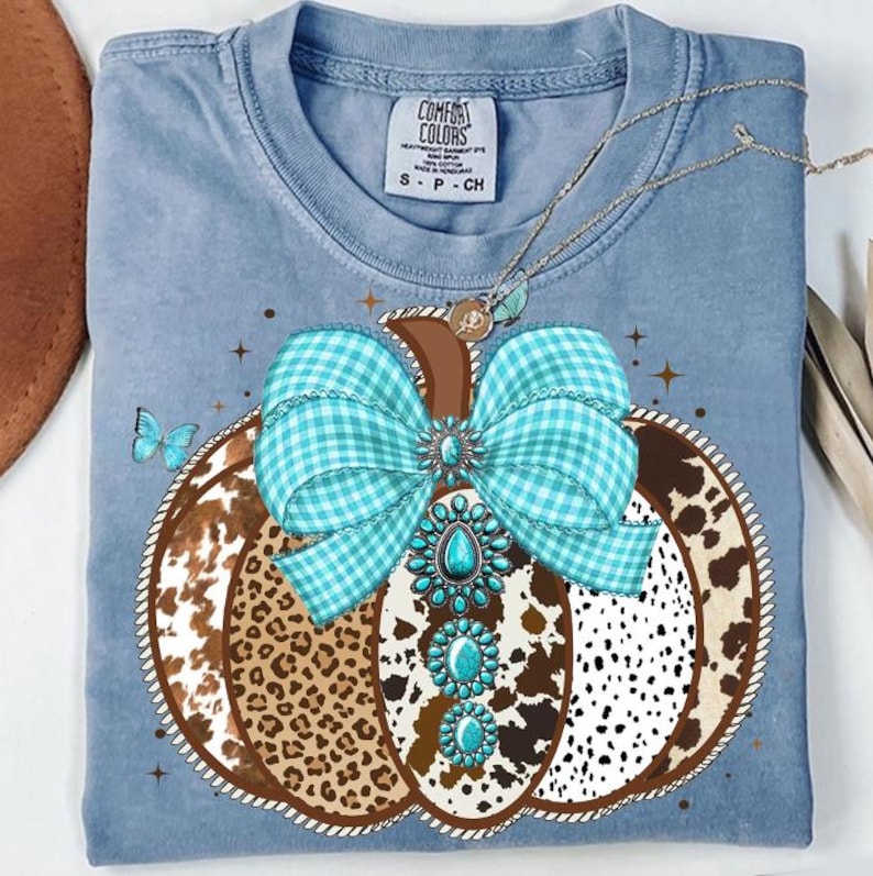 May include: A light blue Comfort Colors t-shirt featuring a pumpkin design. The pumpkin has a brown stem, a turquoise gingham bow, and a turquoise jewel design. The pumpkin is decorated with a cow print and a butterfly.