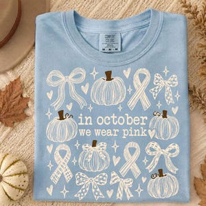 May include: A light blue t-shirt featuring a white graphic print of pumpkins, ribbons, and bows. The text "in october we wear pink" is centered on the chest. The shirt has a crew neckline and a Comfort Colors brand label inside the collar.