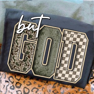 Peut inclure: T-shirt gris foncé avec le mot "GOD" dans un design en lettres majuscules. Les lettres présentent un motif camouflage, vieilli et à carreaux. Le mot "but" est au-dessus du mot "GOD" en écriture blanche.