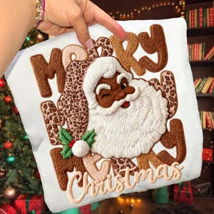May include: White Christmas-themed pillow with an embroidered design. The design features a winking Santa Claus with a white beard and hat, and the words "MERRY CHRISTMAS" in brown and tan colors. The letter "M" and "R" have a leopard print pattern.