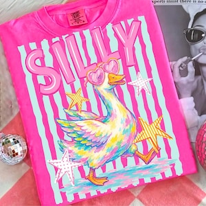 Pode incluir: Uma t-shirt rosa brilhante com a palavra "SILLY" em letras grandes. A t-shirt apresenta um ganso de desenho animado colorido usando óculos de sol em forma de coração e estrelas. O fundo tem listras verticais.