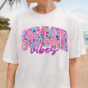 K&ouml;nnte beinhalten: Wei&szlig;es T-Shirt mit einem pink- und blaufarbenen Blumendesign, auf dem "Summer Vibes" steht.