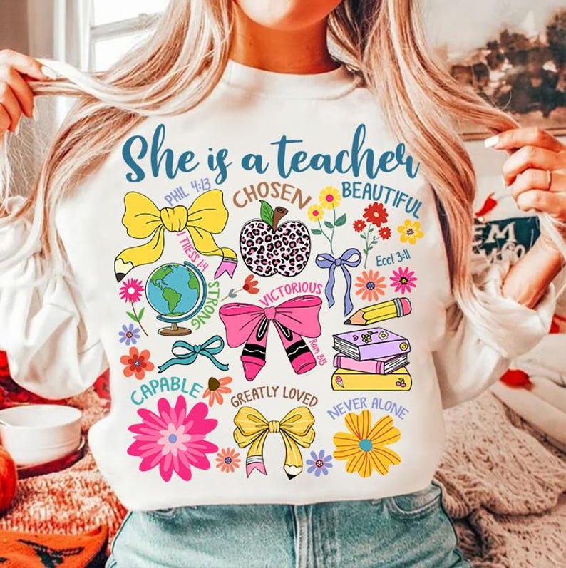 以下が含まれることがあります： ピンクと黒の文字で「She is a teacher」と書かれた白いスウェットシャツ。デザインには、地球儀、リボン、リンゴ、鉛筆、本など、さまざまなカラフルなイラストが描かれています。「Chosen」、「Beautiful」、「Victorious」、「Capable」、「Greatly Loved」、「Never Alone」、「Phil 4:13」、「Thess 1:4」、「Ram 8:13」、「Eccl 3:11」のテキストもデザインに含まれています。