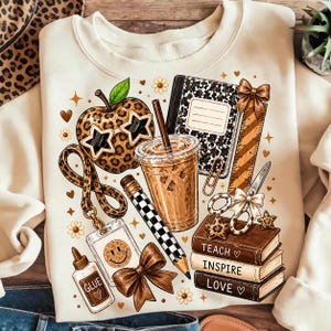Peut inclure: Un sweat-shirt crème avec une impression graphique de fournitures scolaires, incluant une pomme à motif léopard avec des lunettes en forme d'étoile, un café, un carnet, une règle, des ciseaux, un crayon, de la colle et des livres marqués Teach, Inspire, Love.