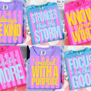 May include: A collection of six crewneck t-shirts featuring vertical stripes and inspirational text. The shirts display phrases including always BE KIND, STRONGER than the STORM, KNOW your WORTH, made for MORE, created WITH A PURPOSE, and FOCUS on the GOOD.