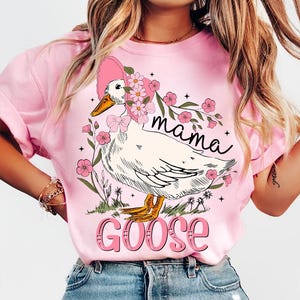 Pu&ograve; includere: Maglietta rosa con una grafica di un'oca che indossa un cappello rosa e fiori. Il testo "mama goose" &egrave; stampato sulla maglietta.